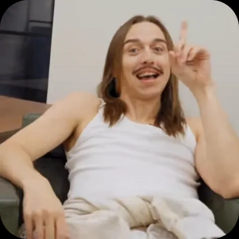 Мужчина в белой майке сидит в кресле, сначала держит руку у лица, потом поднимает палец вверх.