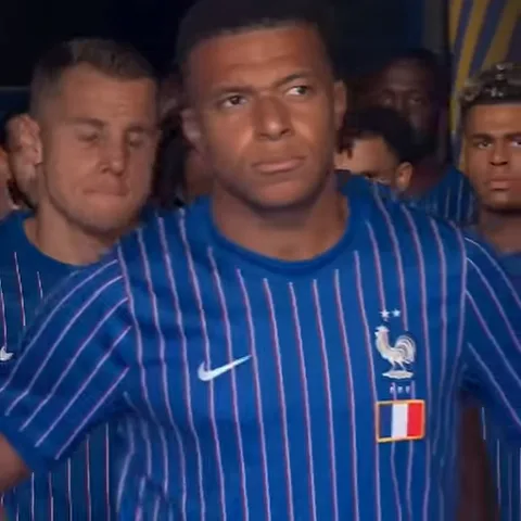 Футболист Килиан Мбаппе в синей полосатой форме сборной Франции поворачивает голову из стороны в сторону, находясь среди других игроков.