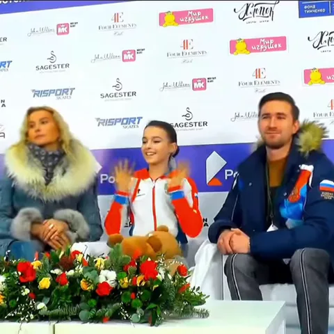 Юная девушка в спортивной куртке с российским гербом сидит за столом с цветами, улыбается и показывает руками сердце.
