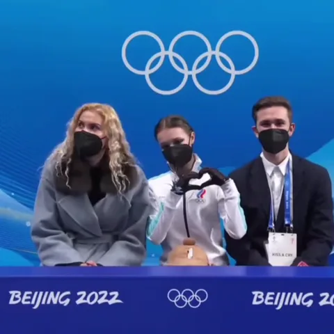 Три человека стоят за барьером с надписью Beijing 2022. В центре девушка в белой куртке и черной маске складывает руки в форме сердца, затем держит медведя.