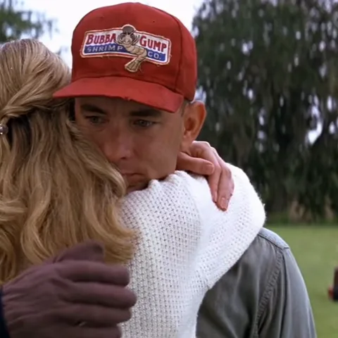 Мужчина в красной бейсболке с надписью «Bubba Gump Shrimp Co.» обнимает женщину, одетую в белый вязаный свитер. На его лице выражение глубокой привязанности и спокойствия.