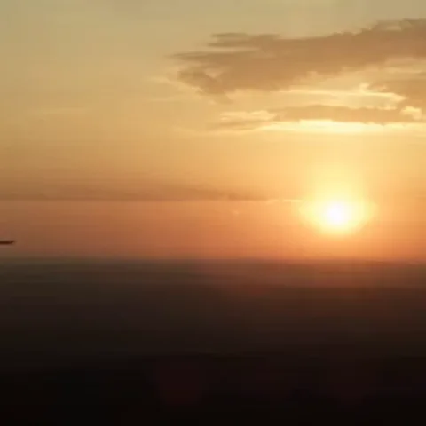 Самолёт летит на фоне оранжевого неба с облаками и ярким солнцем.