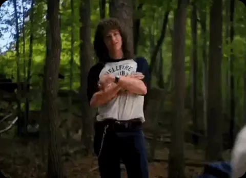 Мужчина с кудрявыми волосами в футболке и джинсах танцует в лесу.