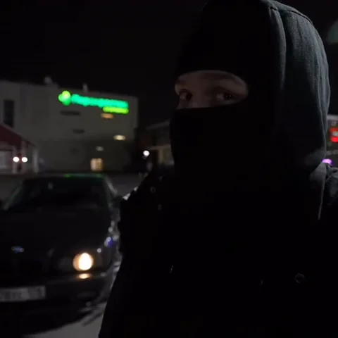 Крупный план лица парня в черной маске, который подмигивает на фоне ночного города с размытыми огнями.