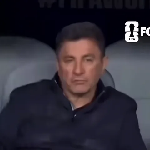Крупный план мужчины с темными волосами в черной куртке, который сидит неподвижно и смотрит в камеру пустым взглядом.