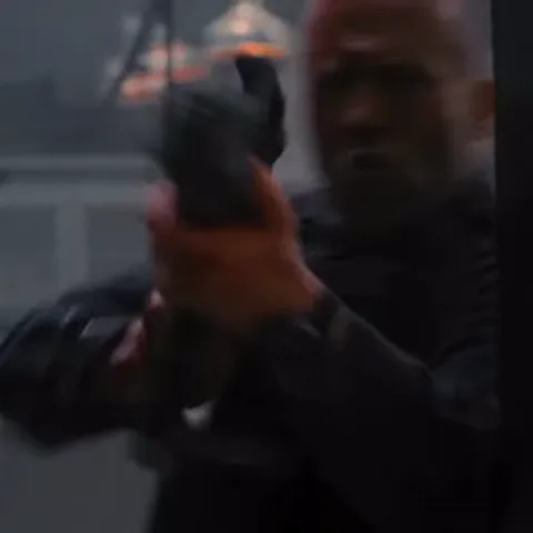 Джейсон Стейтем стреляет из пистолета, из ствола вырывается пламя, на заднем плане броневик.