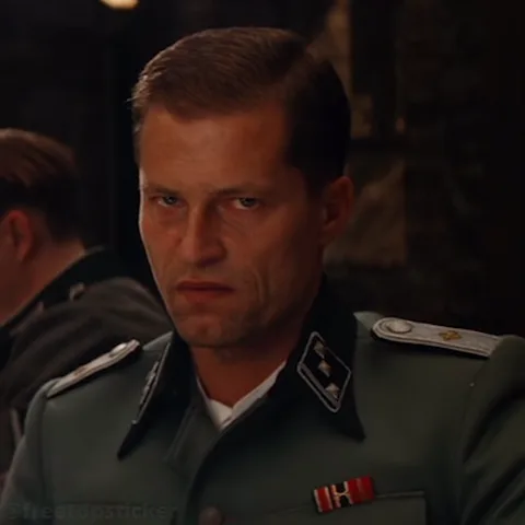 Крупный план мужчины в военной форме, который пристально и холодно смотрит в камеру.