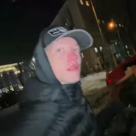 Молодой человек в темной куртке с капюшоном и бейсболке на фоне ночного города делает грустное лицо.
