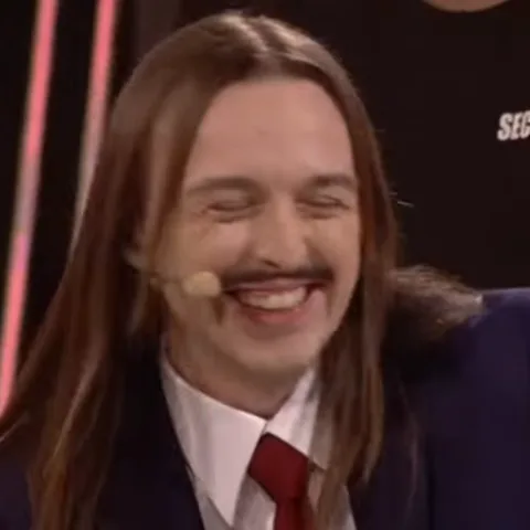 Tommy Cash в костюме с красным галстуком смеется на камеру.