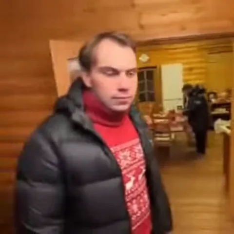 Мужчина в зимней одежде с оленьим принтером на красном фоне стоит внутри бани или деревянного дома.