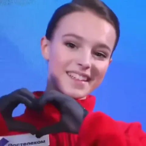 Анна Щербакова в красной куртке делает жест «сердечко» руками в черных перчатках на фоне синей стены.
