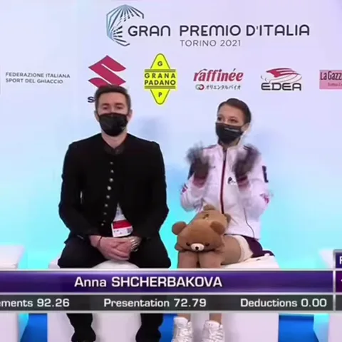 Анна Щербакова в белой куртке и черной маске сидит рядом с тренером, перед ней медведь, она складывает руки в виде сердца.