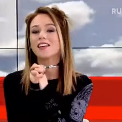 Молодая девушка с длинными светлыми волосами и в черной одежде сидит на фоне экрана с облаками. Она активно жестикулирует рукой, поднося её к шее и касаясь тонкого металлического ожерелья-чокера.