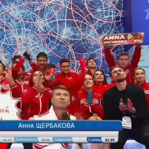 Молодая женщина в красной спортивной куртке улыбается и обнимает большое красное плюшевое сердце. На заднем плане группа людей в похожей форме радостно машет руками.