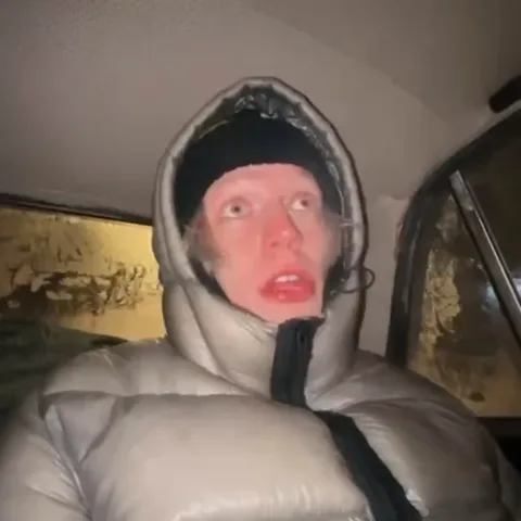 Человек в светлом пуховике с поднятым капюшоном смотрит вверх с открытым ртом и широко распахнутыми глазами, выражая крайнее изумление.