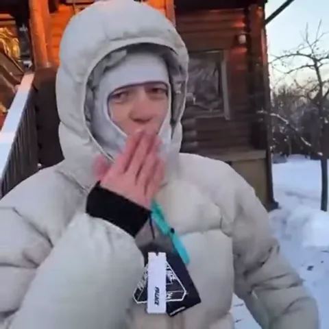 Человек в светлом пуховике и шапке на фоне заснеженного дома жестикулирует рукой и улыбается.