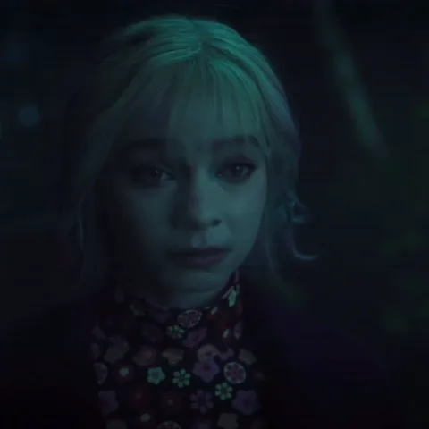 Крупный план Энид Синклер из сериала «Уэнсдей» в тёмной локации с зелёной подсветкой. Девушка с выбритыми висками и цветными прядями активно двигает губами, рассказывая что-то.