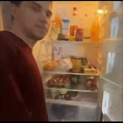 Парень в бордовой футболке смотрит на полки открытого холодильника с продуктами.