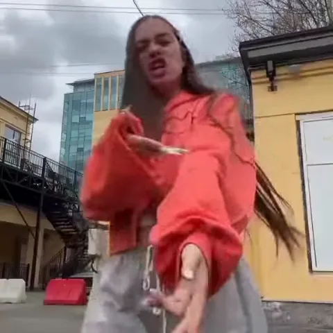 Девушка в оранжевой куртке танцует на фоне желтого здания