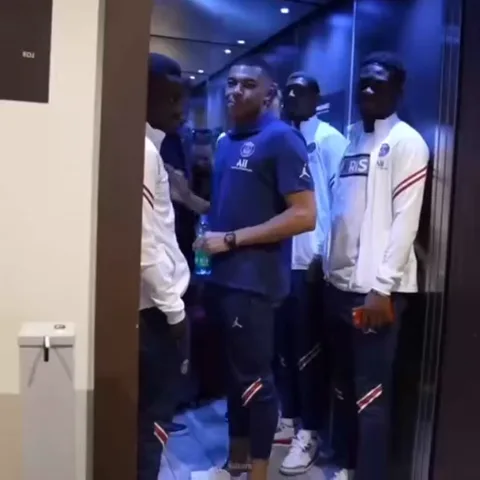 Футболист Килиан Мбаппе в синей форме PSG машет рукой, стоя в лифте среди других игроков.