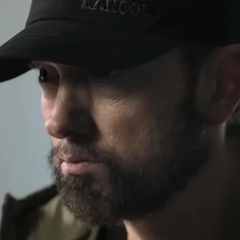Эминем в черной кепке с надписью Kangol смотрит в сторону с задумчивым выражением лица.