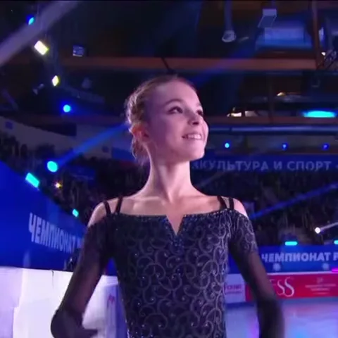 Анна Щербакова в черном платье с блеском на льду делает жест «сердечко» из рук и улыбается.