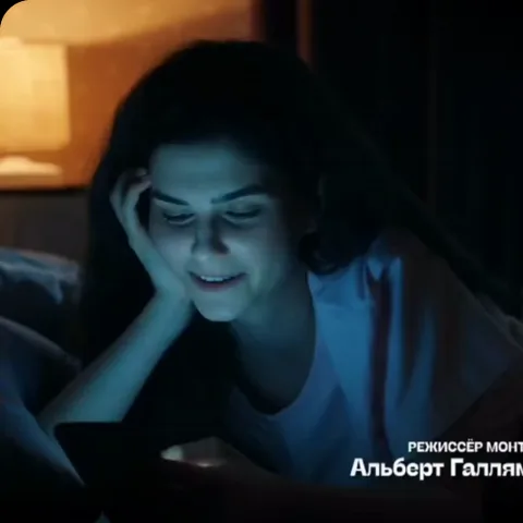 Брюнетка в белой футболке лежит в темноте, опираясь на руку, и нежно улыбается, глядя вниз в экран телефона.