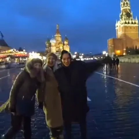 Ночная сцена на Красной площади: три подруги в тёплой одежде смеются и позируют, одна размахивает рукой, на заднем плане подсвеченные исторические здания.