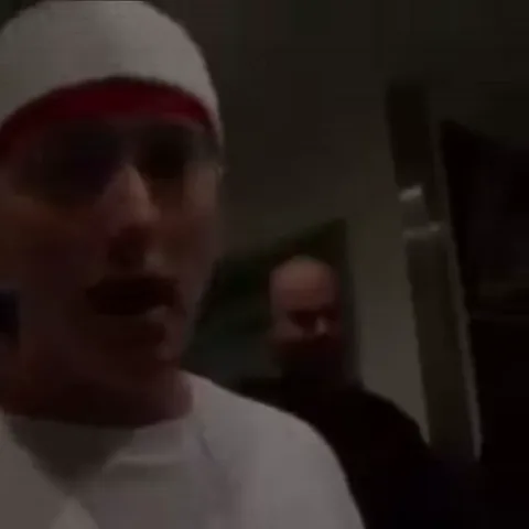 Эминем в белой одежде и шапке с красной полосой активно жестикулирует перед камерой.