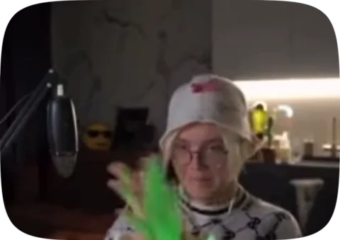 Молодой человек с блондинистыми волосами, в очках и белой шапкеbucket hat, улыбается и поднимает руку с надетой ярко-зеленой перчаткой в виде звериной лапы. На заднем плане виден микрофон.
