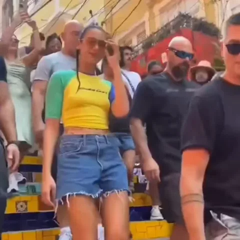 Женщина с темными волосами, собранными в хвост, в желто-зеленой футболке и джинсовых шортах, идет вниз по лестнице и поднимает руку, чтобы помахать. Вокруг нее мужчины в черной одежде (охрана) и толпа людей на фоне.