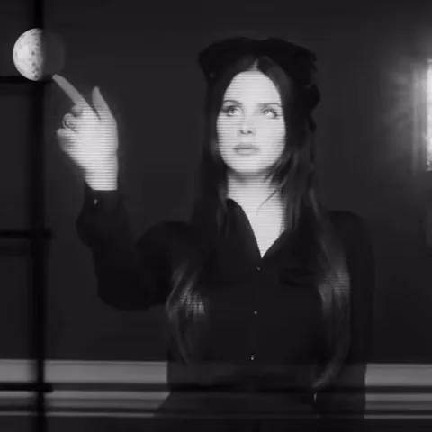 Лана Дель Рей в чёрной рубашке смотрит вверх и указывает пальцем на луну на тёмном фоне.