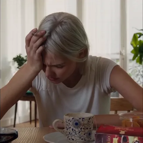 Светловолосая девушка в белой футболке сидит за деревянным столом, прижимает руку к голове и курит сигарету, перед ней кружка с чаем.