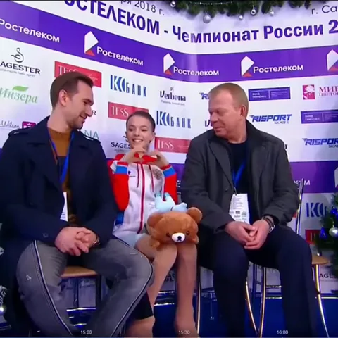 Анна Щербакова в спортивной куртке показывает руками сердце на фоне баннера с логотипами спонсоров.