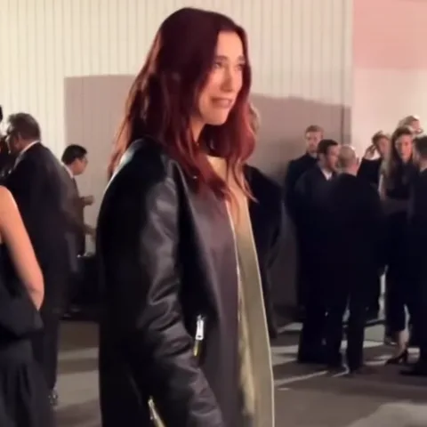 Dua Lipa позирует на мероприятии, одетая в черную кожаную куртку и золотое ожерелье.