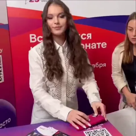 Девушка в белой рубашке на фоне красного баннера делает жест «сердце» из рук и улыбается.