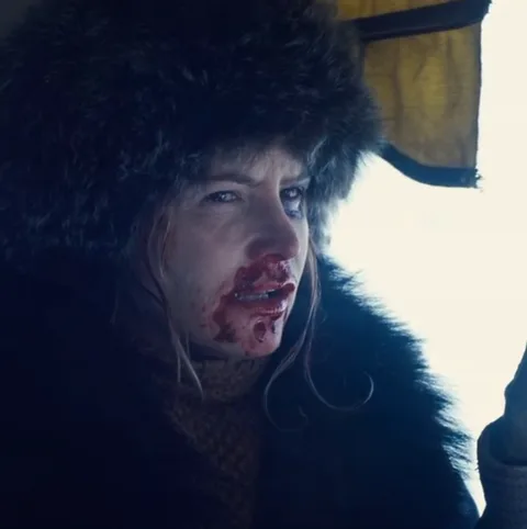 Крупный план женщины в меховой шапке и шубе. У неё кровь на лице, она смотрит в сторону и постепенно начинает улыбаться.