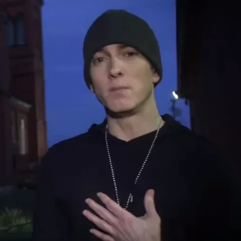 Эминем в черной одежде и шапке говорит и разводит руками на фоне вечернего неба.