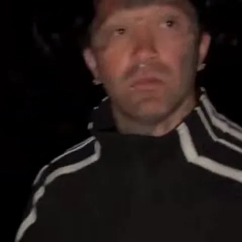 Мужчина с татуировками на лице и в черной куртке Adidas пристально смотрит в камеру, а затем его лицо крупным планом заполняет кадр.