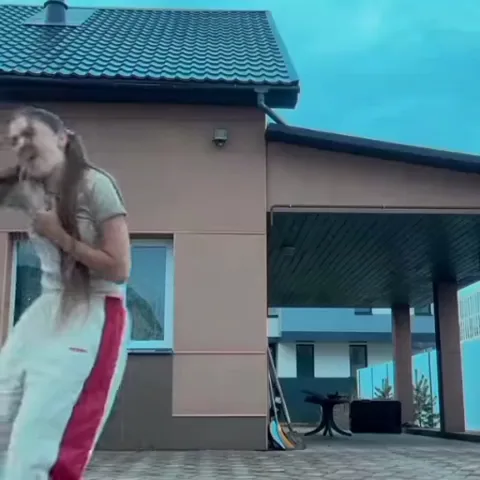Девушка в белом спортивном костюме машет рукой, а затем подзывает к себя обеими ладонями на фоне дома.