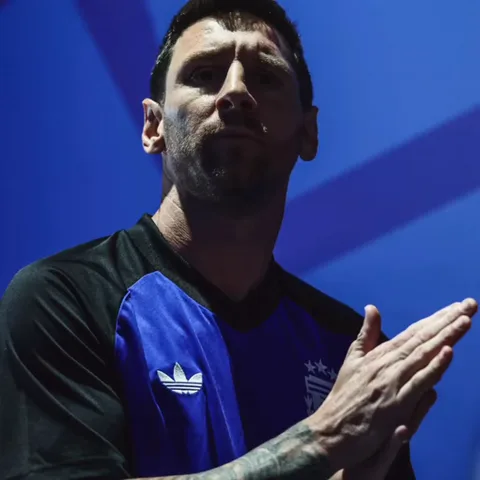 Футболист Лионель Месси в чёрно-розовой футболке с логотипом Adidas стоит на синем фоне и хлопает в ладоши, глядя прямо перед собой.