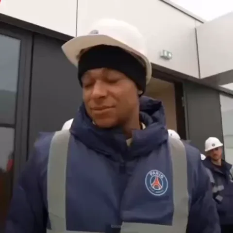 Футболист Килиан Мбаппе в белой каске и синей куртке с логотипом ПСЖ смотрит в сторону с довольным выражением лица.