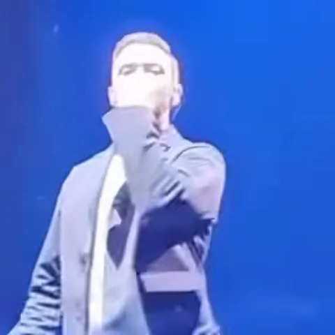 Джастин Тимберлейк в сером костюме на фоне синего света делает пальцами знак «ОК».