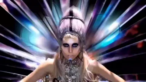 Lady Gaga в серебристом платье и с крыльями, стоящая на фоне абстрактных лучей света.