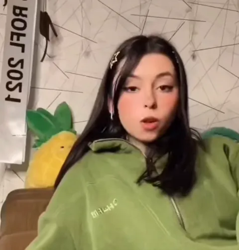 Брюнетка в зелёной олимпийке с надписью CHILL на груди. Она устало прикрывает глаза рукой, демонстрируя жест разочарования или смущения, после чего заваливается назад.
