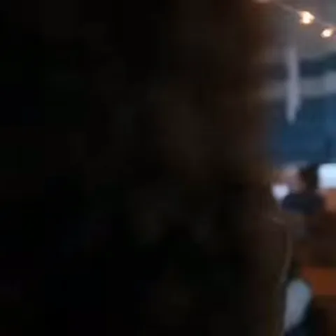 Подросток с темными волосами в коричневом пиджаке и голубом свитере стоит перед блестящими гирляндами.