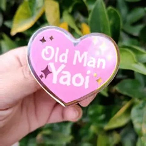 Крупный план руки, держащей розовый брелок в форме сердца с надписью Old Man Yaoi и искрами, на фоне зеленых листьев.
