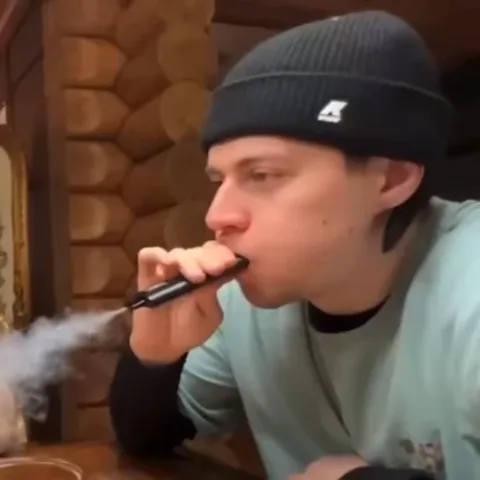 Человек в черной шапке затягивается вейпом и выдыхает дым ноздрями.