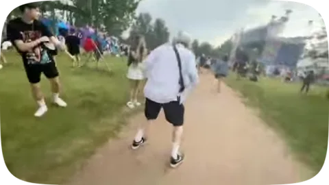 Молодой человек с выбеленными волосами, одетый в белую футболку и темные шорты, активно двигается и пританцовывает на фоне толпы людей и деревьев.