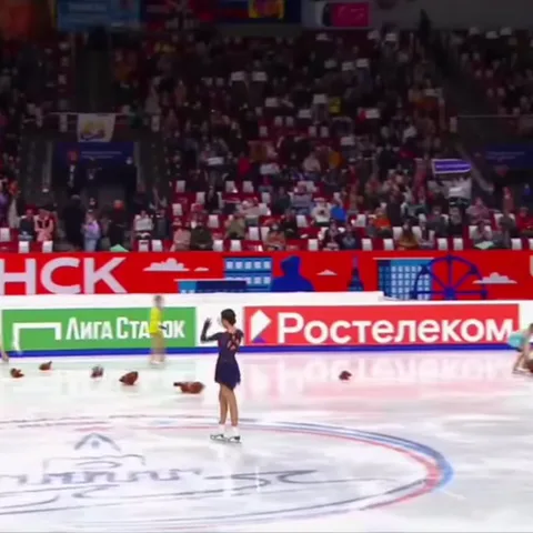 Фигуристка Анна Щербакова в синем платье на ледовой арене, вокруг разбросаны плюшевые игрушки, она поднимает руку, чтобы помахать публике.
